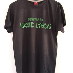Könnte beinhalten: Schwarzes T-Shirt mit dem Text "Directed by DAVID LYNCH" in grünen Buchstaben. Das T-Shirt hängt an einem Holzbügel vor weißem Hintergrund. Das T-Shirt hat einen Rundhalsausschnitt und kurze Ärmel.