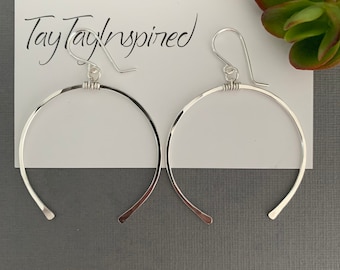 May include: A pair of silver hoop earrings with a hammered texture. The earrings are on a white card with the text "TayTayInspired" in black script. A green succulent plant is in the background.