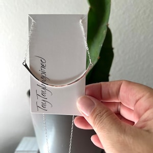 May include: A silver-colored necklace with a curved bar pendant. The necklace is displayed on a white card with the text "TayTaylorInspired" in a handwritten font. The necklace hangs from a delicate chain.