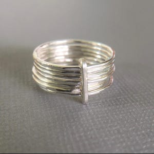 May include: A silver ring with a unique design. The ring features multiple bands held together by a vertical bar. The bands have a textured surface, giving the ring a handcrafted appearance. The ring is set against a gray background.