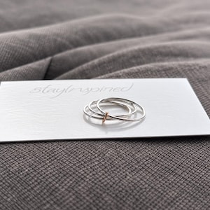 May include: A set of three thin stacking rings resting on a white card with the text stayinspired printed in gray. Two rings are silver-toned, and one ring features a small rose gold-toned accent piece. The rings are smooth and circular.