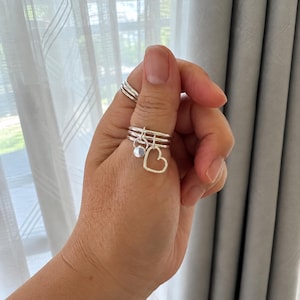 May include: A set of three silver-colored rings worn on a thumb. The middle ring features two dangling charms: one small circular disc and one open-frame heart shape. The rings have a smooth, polished finish and a simple, thin band design.