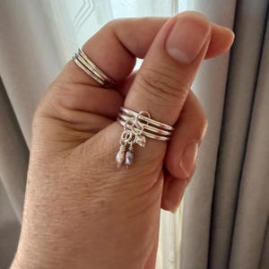 May include: A set of three stacked silver rings worn on a thumb, featuring three dangling charms. Two charms are small, teardrop-shaped iridescent pearls in light pink and lavender, and the third charm is a small, polished silver heart shape.