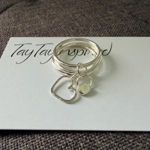May include: A silver ring set consisting of three thin bands stacked together. Two small charms hang from the rings: one is a simple open heart outline and the other is a small circular disc. The ring rests on a white card with TayTayInspired text.