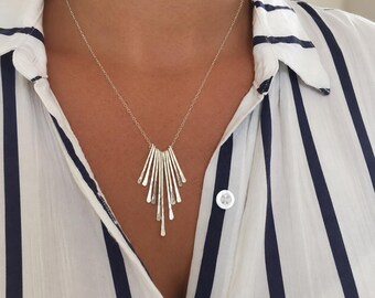 May include: A silver-toned necklace featuring a pendant composed of multiple thin, hammered metal bars of varying lengths arranged in a fan-like, sunburst pattern, suspended from a delicate, fine-link silver-toned chain.
