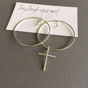 May include: A pair of silver hoop earrings with a cross pendant. The earrings are displayed on a white card with the handwritten text "TayTayInspired". The jewelry is set against a gray background.