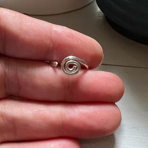 May include: A silver-colored metal ring featuring a prominent spiral design at the center. The band is open-ended, allowing for adjustment, and the surface of the metal has a smooth, polished finish that reflects light across its curved shape.
