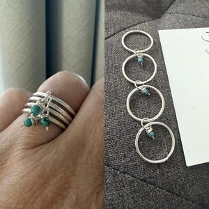 May include: A silver ring with four bands, adorned with three turquoise beads. Next to it, four silver hoop rings, each with a turquoise bead, are displayed. The rings are on a gray surface.