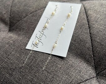 Puede incluir: Un par de pendientes largos de cadena plateada con pequeñas perlas de agua dulce blancas e irregulares espaciadas a lo largo de cada cadena. Los pendientes se presentan en una tarjeta rectangular blanca con el texto TayloredJewelry.