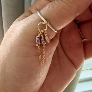 May include: A silver ring with a gold chain and three dangling, irregularly shaped, purple and pink pearls. The ring is worn on a finger, with other silver rings visible in the background.