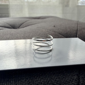 May include: A silver-colored ring featuring a multi-coil spiral design with a hammered, textured finish. The band wraps around itself three times, creating a layered appearance with a polished, reflective surface that catches the light.