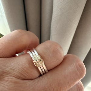 May include: A stackable ring set consisting of three silver-colored bands with a hammered texture and two thinner, smooth rose gold-colored bands intertwined with the silver rings, creating a layered appearance on the finger.