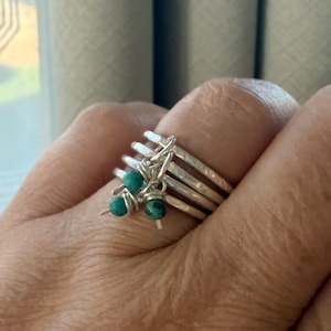 May include: A stackable silver ring set with turquoise-colored beads. The rings have a hammered texture and are connected with silver wire. The turquoise beads are small and round, adding a pop of color to the design. The rings are on a person's finger.