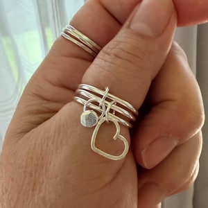 May include: A set of silver rings on a finger, with a heart and a small charm dangling from the rings. The rings are stacked together, and the heart charm is open. The rings are on a finger.