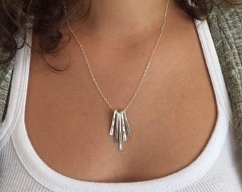 May include: A silver-toned necklace featuring a pendant composed of five thin, vertically hanging, hammered metal bars of varying lengths. The pendant is suspended from a delicate, fine-link silver-toned chain that rests against the collarbone area.