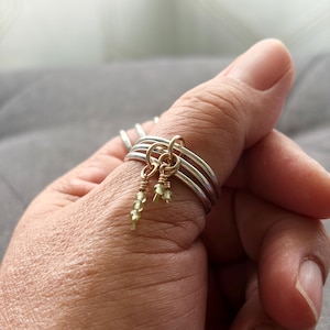 May include: A stack of four silver rings worn on a thumb. Two small gold jump rings are attached to the silver bands, each featuring a dangling charm made of tiny, pale green faceted beads wrapped with thin gold wire for a delicate appearance.