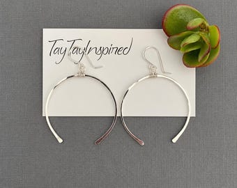 May include: A pair of silver hoop earrings with a crescent shape and wire-wrapped detail at the top, attached to a white display card with the text TayTayInspired printed in black, accompanied by a small green succulent plant.