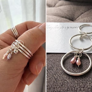 May include: Silver rings with dangling charms and small pink pearls. One ring is on a finger, and the other rings are displayed on a white card. The rings are connected in a chain.