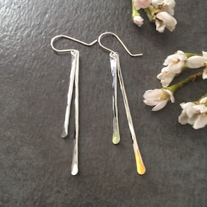 May include: A pair of silver dangle earrings with a hammered texture. Each earring has a long, slender bar with a teardrop-shaped end. The earrings are displayed on a dark gray surface, with white flowers in the background.