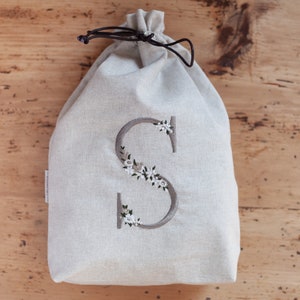 May include: A beige drawstring bag with a large embroidered letter "S" adorned with white and green floral accents. The bag is made of a textured fabric and has a dark brown drawstring closure. The bag is sitting on a wooden surface.