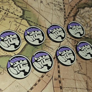 Può includere: Una collezione di otto gettoni rotondi in bianco e nero con un design viola. Ogni gettone presenta un teschio stilizzato con un copricapo viola. I gettoni sono disposti su una mappa vintage.