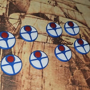 May include: A collection of eight circular, hand-painted game pieces. Each piece features a white circle with a blue cross design and a red center. The pieces are arranged on a brown, textured surface.