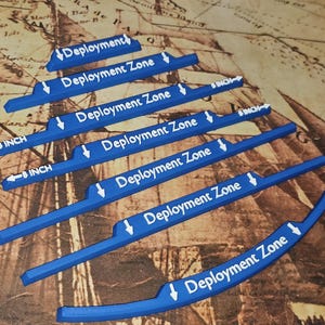 Op de afbeelding: Blauwe plastic spelstukken met de tekst "Deployment Zone" en "INCH" in het wit. De stukken zijn van verschillende lengtes, met pijlen die de richting aangeven. De achtergrond is een kaart.
