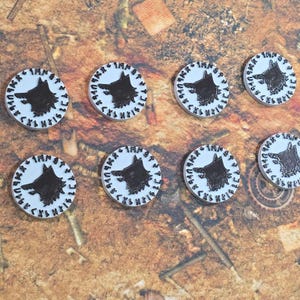 May include: Eight white, circular game tokens with a black wolf head design in the center. Each token has a black border with runes. The tokens are arranged on a brown, textured surface.