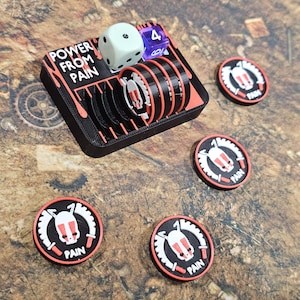May include: A black and red game piece holder with the text "POWER FROM PAIN" and several black and red game tokens. A white die with black dots and a purple die with a 4 are also present. The tokens have a white and red design with the word "PAIN".