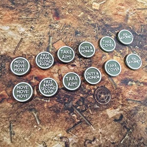 Op de afbeelding: Ronde spel fiches met commando's zoals "MOVE!", "TAKE AIM!" en "DUTY & HONOR" erop gedrukt. De fiches zijn olijfgroen met witte tekst en een zilveren rand, op een bruin oppervlak.