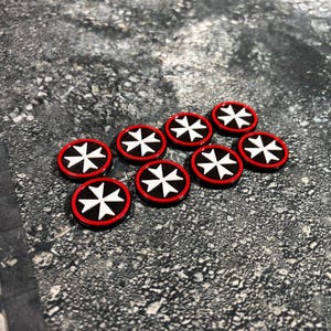 May include: A collection of eight circular tokens with a red border, black center, and a white Maltese cross design. The tokens are arranged on a textured gray surface, suitable for tabletop gaming or crafting projects.