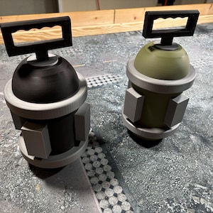 May include: Two 3D-printed grenade-like objects. One is black, the other olive green. Each has a cylindrical base with rectangular blocks, a rounded top, and a black handle. They are displayed on a textured surface.