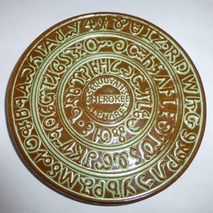 May include: A round brown ceramic plate featuring a light green glazed relief of the Cherokee alphabet arranged in concentric circles. The center of the plate displays the text SEQUOYAH'S CHEROKEE ALPHABET in a circular embossed design.