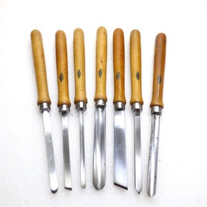 May include: A set of seven woodturning chisels with light brown wooden handles and polished steel blades. Each tool features a metal ferrule connecting the handle to the blade, and a small oval label is attached to the side of each wooden handle.
