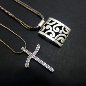 May include: Two silver-toned necklaces. One features a rectangular pendant with an openwork scroll pattern on a braided chain. The second is a cross pendant encrusted with small clear stones on a thin snake chain, both with a metallic finish.