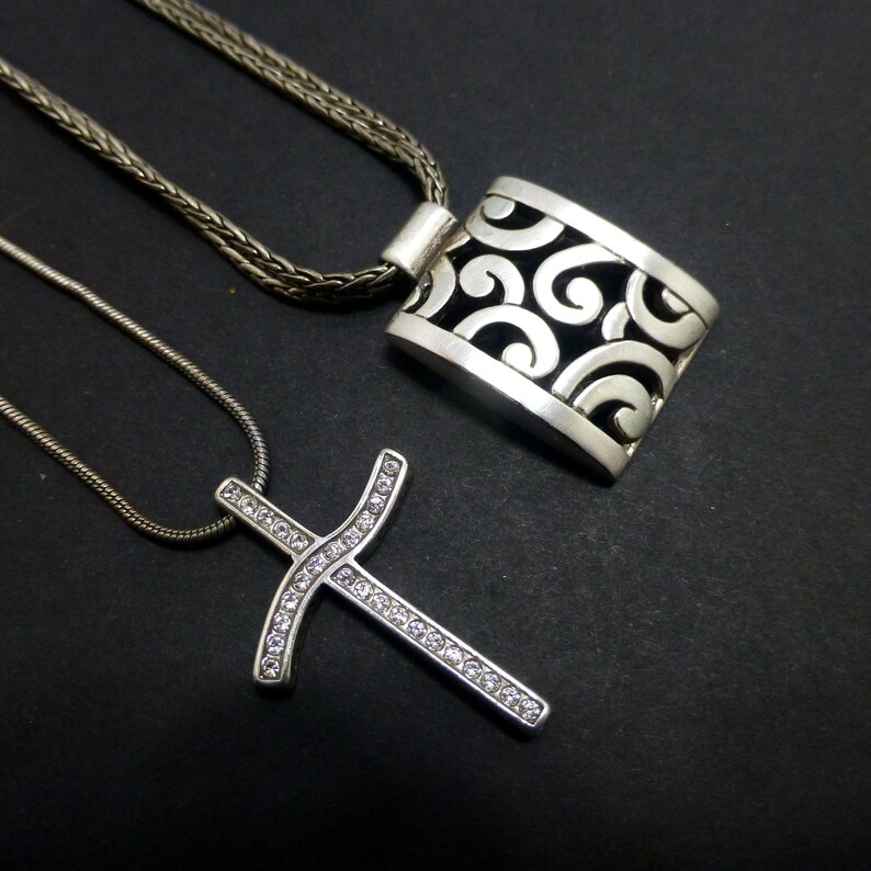 May include: Two silver-toned necklaces. One features a rectangular pendant with an openwork scroll pattern on a braided chain. The second is a cross pendant encrusted with small clear stones on a thin snake chain, both with a metallic finish.