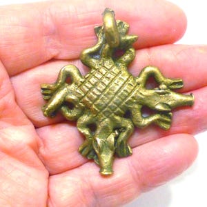 May include: A brass-toned metal pendant featuring a central square with a cross-hatched grid pattern, surrounded by four stylized animal figures with elongated snouts and curved limbs, topped with a small circular loop for attachment.