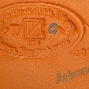 K&ouml;nnte beinhalten: Nahaufnahme eines terrakottafarbenen Keramiksockels mit gepr&auml;gtem Text und Designelementen. Der Text enth&auml;lt "MacKenzie-Childs, Ltd.", "Aurora, New York 13026" und "Aalsmeer". Das Design zeigt ein zentrales Quadrat mit einem "W" und einem "C"-Symbol.