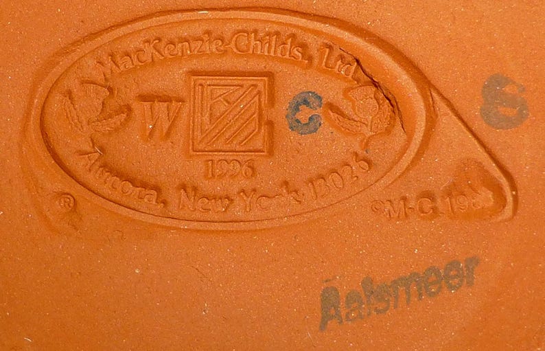 K&ouml;nnte beinhalten: Nahaufnahme eines terrakottafarbenen Keramiksockels mit gepr&auml;gtem Text und Designelementen. Der Text enth&auml;lt "MacKenzie-Childs, Ltd.", "Aurora, New York 13026" und "Aalsmeer". Das Design zeigt ein zentrales Quadrat mit einem "W" und einem "C"-Symbol.
