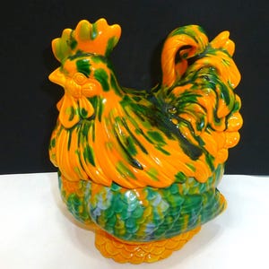 May include: A ceramic rooster-shaped jar featuring a vibrant marbled glaze in shades of bright orange and deep forest green. The lid forms the head and neck of the rooster, while the base displays a textured, scale-like pattern in green and orange.
