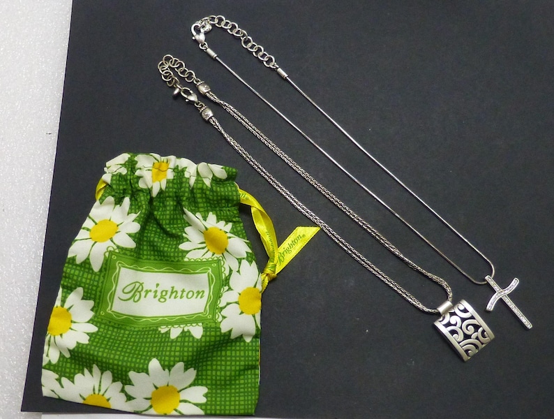 May include: Two silver-tone necklaces with lobster claw clasps and extender chains. One necklace features a rectangular pendant with an openwork scroll pattern, and the other features a cross pendant. A green drawstring pouch with white daisy print.