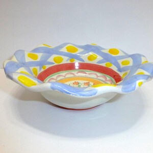 K&ouml;nnte beinhalten: Dekorative Keramikschale mit gewelltem Rand. Die Schale hat einen wei&szlig;en Boden mit farbenfrohem Innendesign. Der Rand ist mit blauen und gelben Mustern bemalt. Das Innere hat rote, orangefarbene und florale Designs.