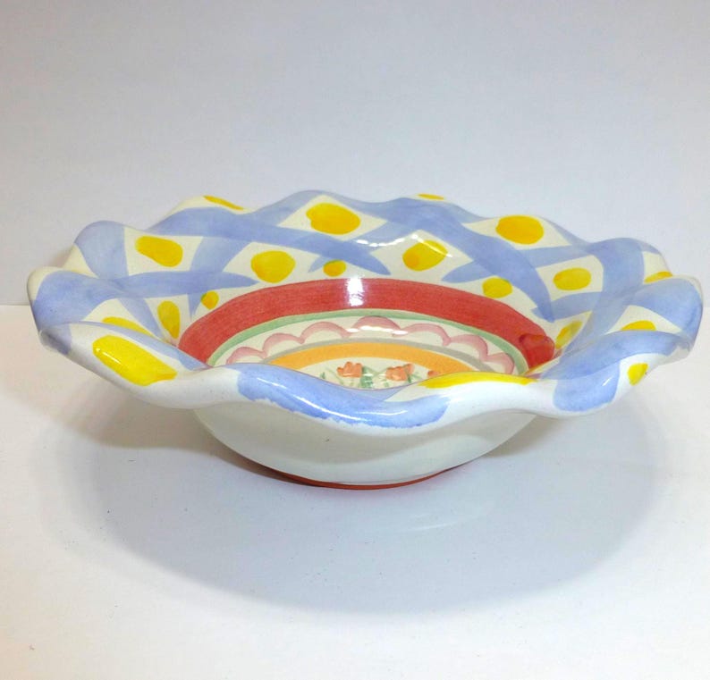 K&ouml;nnte beinhalten: Dekorative Keramikschale mit gewelltem Rand. Die Schale hat einen wei&szlig;en Boden mit farbenfrohem Innendesign. Der Rand ist mit blauen und gelben Mustern bemalt. Das Innere hat rote, orangefarbene und florale Designs.