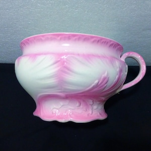 May include: A decorative, vintage-style ceramic bowl with a handle, featuring a white and pink floral design. The bowl has a scalloped base and a curved rim. The handle is a simple, rounded shape. The bowl is likely used for decorative purposes.
