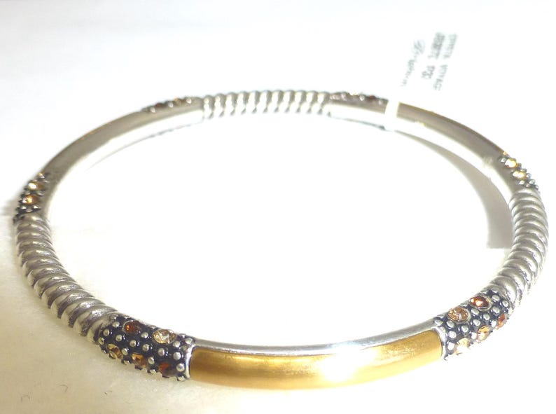 Puede incluir: Pulsera de brazalete plateada con una secci&oacute;n central dorada. La pulsera presenta un dise&ntilde;o de cuerda trenzada en los laterales y est&aacute; adornada con peque&ntilde;as piedras brillantes de color marr&oacute;n y dorado. Una etiqueta est&aacute; adjunta.