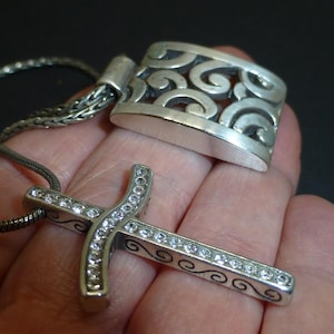 May include: A silver-toned cross pendant featuring a row of clear, sparkling stones embedded along the center, accented with black scrollwork patterns on the sides. The pendant is attached to a thin, flexible silver-toned chain necklace.