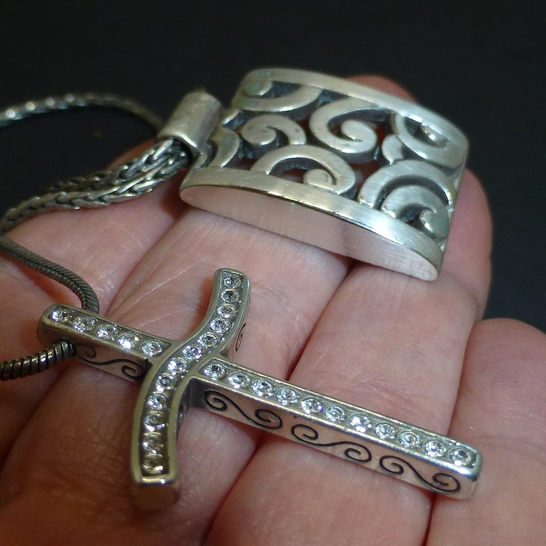 May include: A silver-toned cross pendant featuring a row of clear, sparkling stones embedded along the center, accented with black scrollwork patterns on the sides. The pendant is attached to a thin, flexible silver-toned chain necklace.