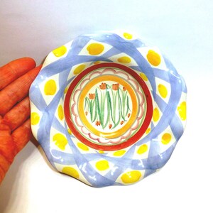 K&ouml;nnte beinhalten: Ein dekorativer Keramikteller mit gewelltem Rand. Der Teller hat ein blau-wei&szlig;es Gittermuster mit gelben Punkten. Die Mitte hat ein Blumenmuster mit orangefarbenen Blumen und gr&uuml;nen Stielen.