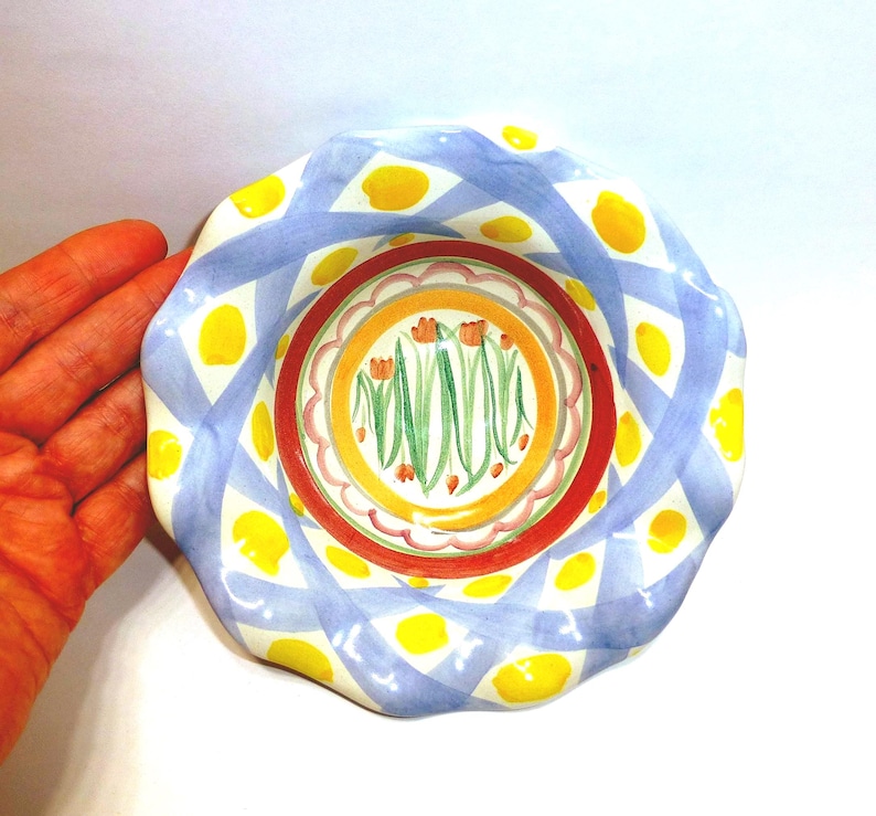 K&ouml;nnte beinhalten: Ein dekorativer Keramikteller mit gewelltem Rand. Der Teller hat ein blau-wei&szlig;es Gittermuster mit gelben Punkten. Die Mitte hat ein Blumenmuster mit orangefarbenen Blumen und gr&uuml;nen Stielen.