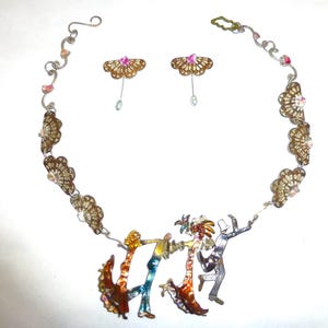 May include: A necklace and earring set featuring a central pendant of four dancing figures in metallic tones. The necklace chain includes fan-shaped charms and small crystals. The matching earrings are fan-shaped with a pink center stone and a dangling bead.